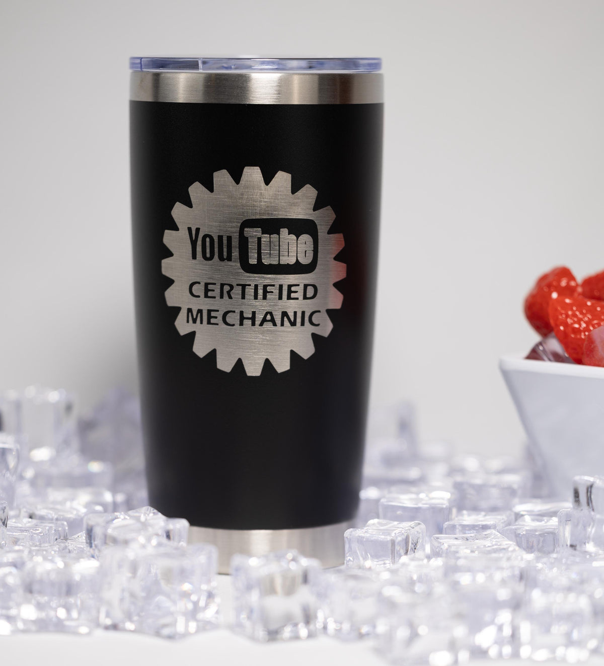 A black tumbler with a silver lid, featuring a logo that says "YouTube Certified Mechanic" on it. The tumbler is surrounded by ice cubes and strawberries.