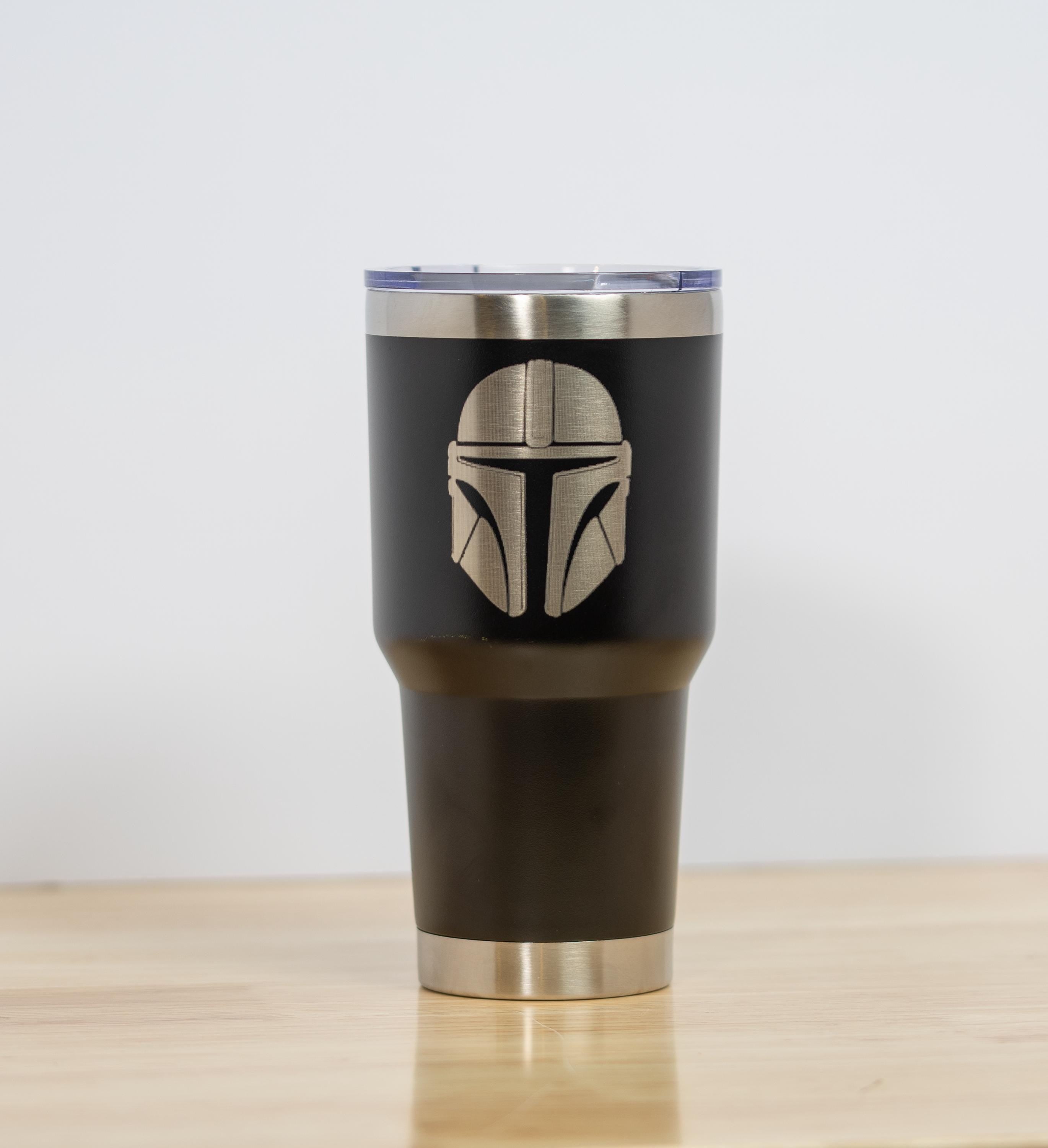 May include: A black insulated tumbler with a silver rim and lid. The tumbler features a detailed silver engraving of a Mandalorian helmet. The tumbler is designed to hold approximately 30 ounces.