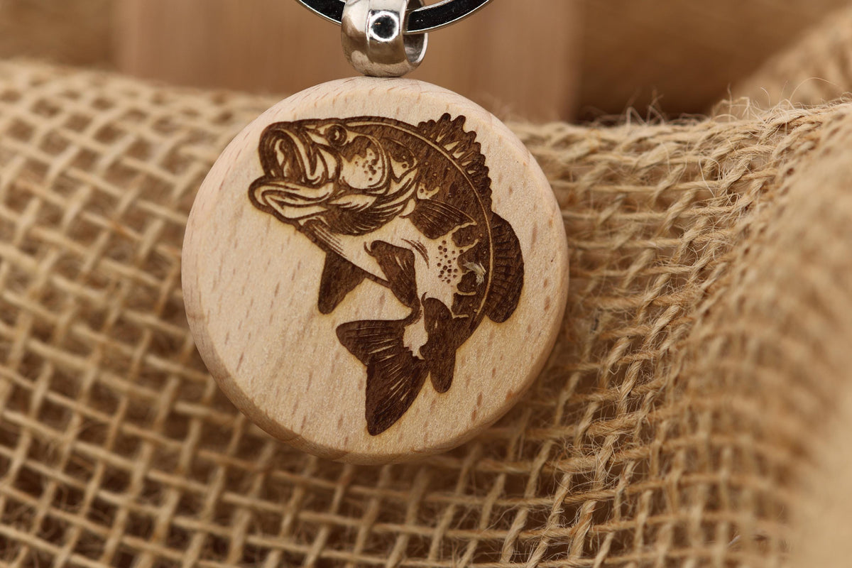 May include: A round wooden pendant with a detailed engraving of a fish. The fish is brown and appears to be a bass, with its mouth open and fins extended. The pendant is attached to a black cord with a silver-colored metal clasp. The background is a textured, tan fabric.