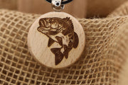 May include: A round wooden pendant with a detailed engraving of a fish. The fish is brown and appears to be a bass, with its mouth open and fins extended. The pendant is attached to a black cord with a silver-colored metal clasp. The background is a textured, tan fabric.