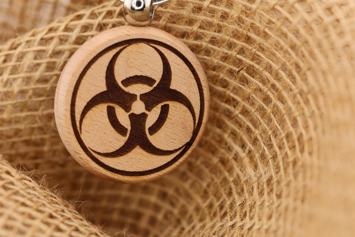 May include: A round wooden pendant with a biohazard symbol engraved in dark brown. The symbol is a circle with three radiating arms. The pendant hangs from a silver-colored chain. The background is a close-up of a rough, tan burlap fabric.