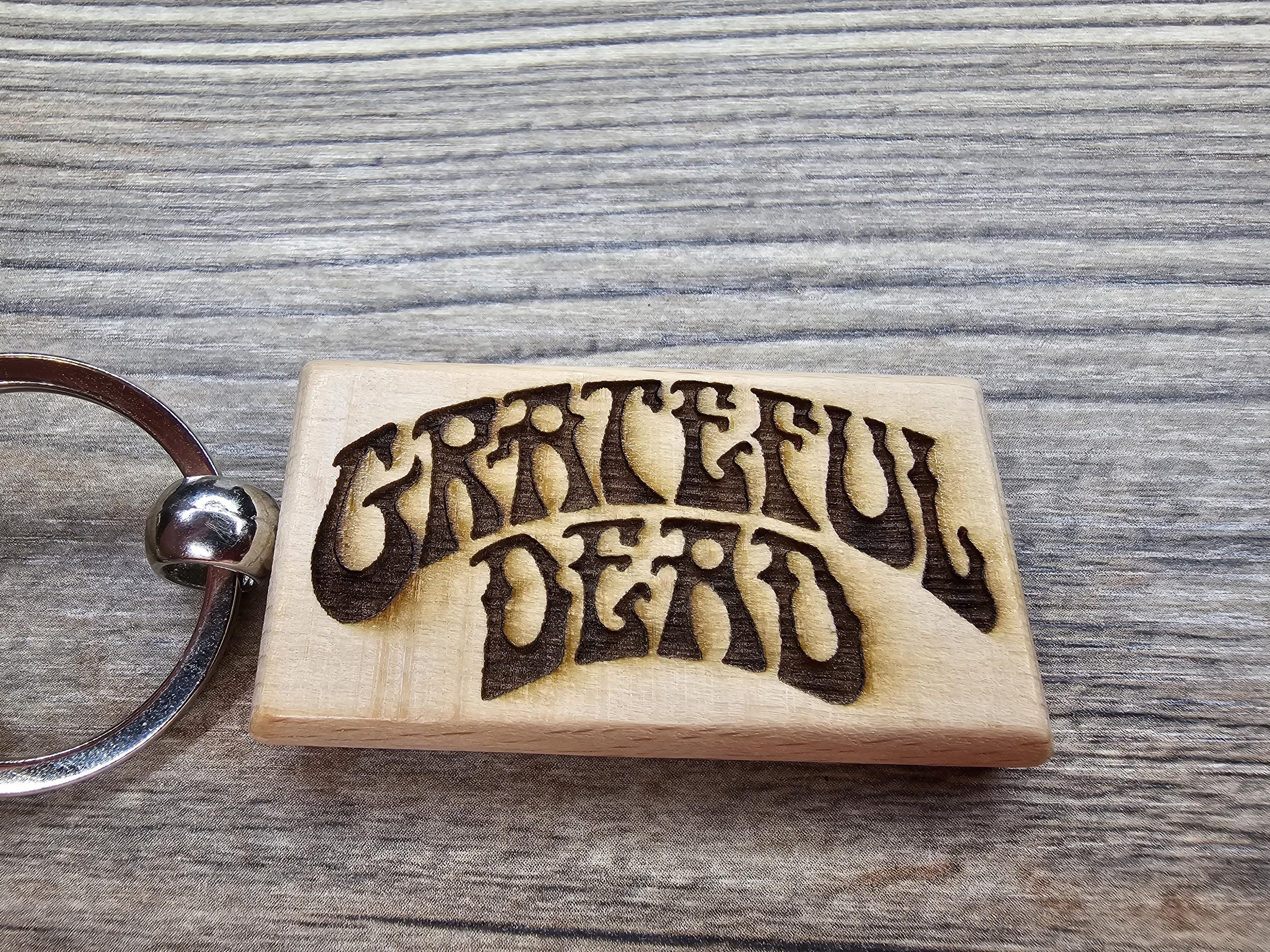 May include: A wooden keychain featuring the words 'Grateful Dead' in a stylized, arched font. The text is dark brown, burned into the light-colored wood. The keychain has a silver metal ring attached to the left side. The wood has a rectangular shape with rounded corners.