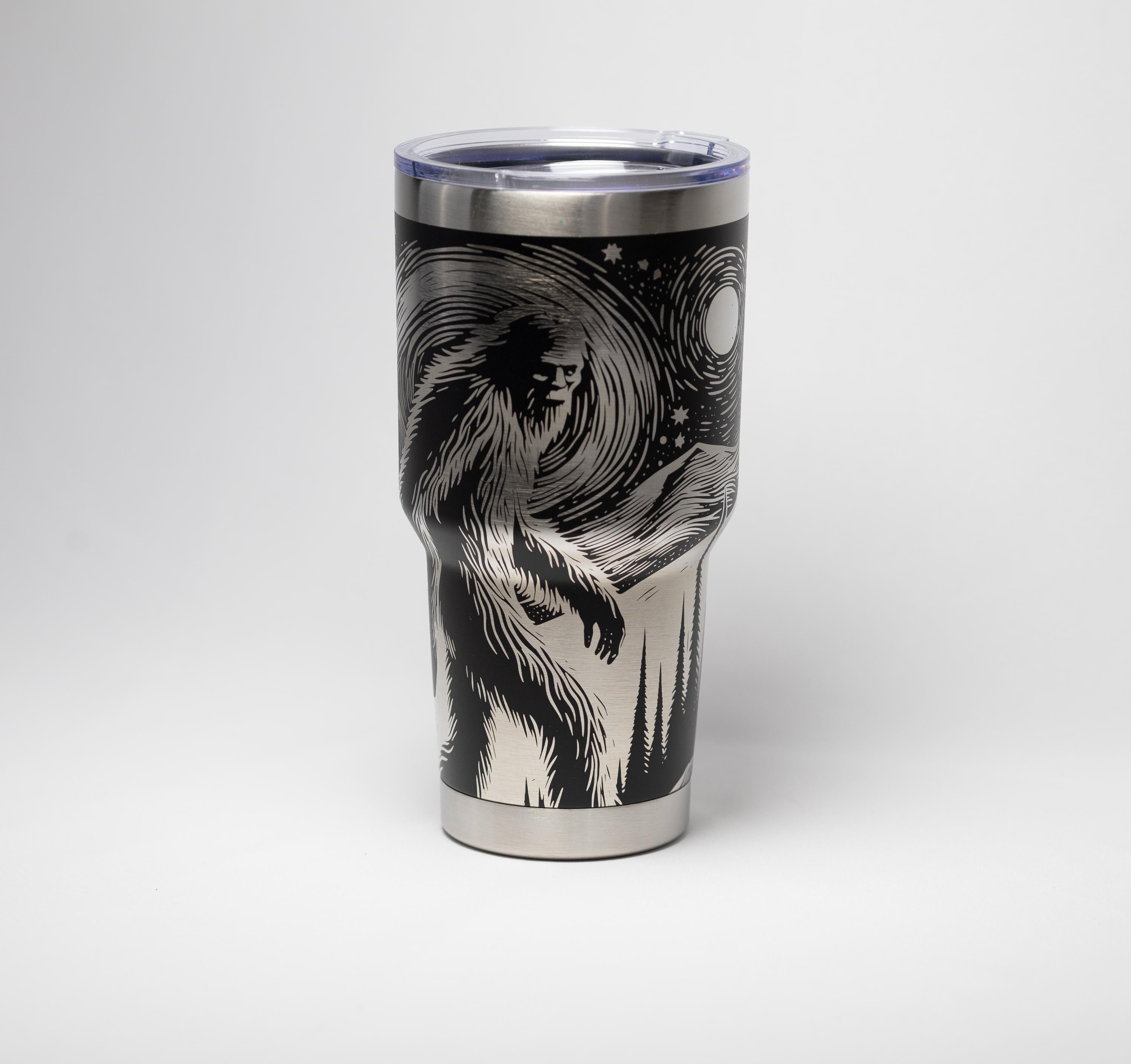 May include: Stainless steel tumbler with a black and white graphic of Bigfoot in a forest scene under a starry night sky. The tumbler has a silver rim and a clear plastic lid. The design wraps around the insulated travel mug.