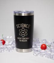 A black insulated tumbler with a silver lid, featuring a humorous text that reads "SCIENCE DOESN'T CARE WHAT YOU BELIEVE". The tumbler is surrounded by ice cubes and a cherry, creating a playful and whimsical scene.