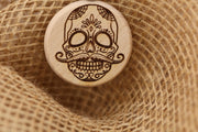 May include: A round wooden pendant with a detailed sugar skull design. The skull features large eye sockets, a mustache, and intricate patterns. The pendant is light brown with dark brown etched details, and is set against a textured, tan background.