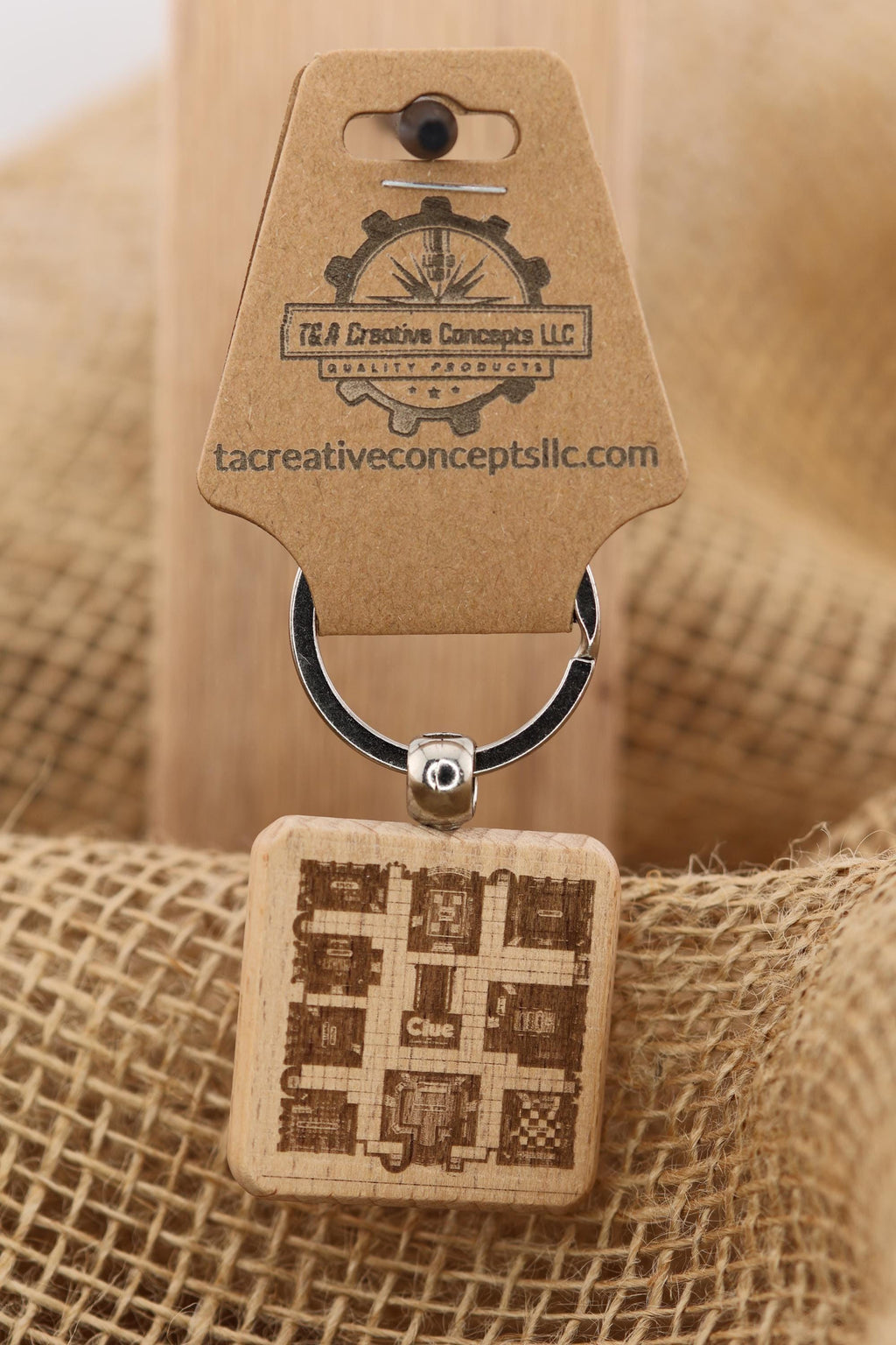 May include: A wooden Clue game keychain. The keychain features a square wooden charm engraved with a map of the game board, the word 'Clue', and the word 'TUS'. The charm is attached to a metal ring and a brown tag with the logo of T&A Creative Concepts LLC and the website tacreativeconceptsllc.com.
