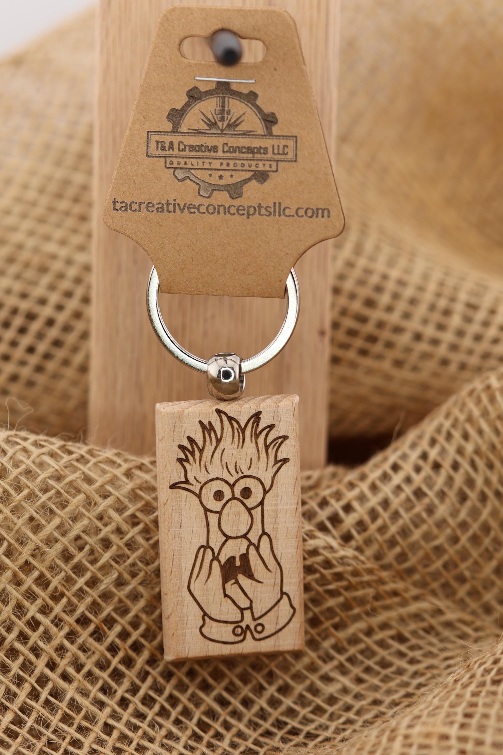 May include: A wooden keychain featuring a cartoon character engraved on a rectangular block. The character has spiky hair, round glasses, and is holding something in its hands. The keychain has a silver ring and a brown tag with the text 'T&A Creative Concepts LLC QUALITY PRODUCTS' and 'tacreativeconceptsllc.com'.